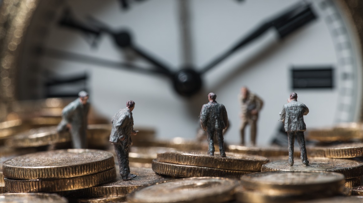 Makro fotografija malih figurica ljudi koje stoje na gomili zlatnih novčića, sa velikim satom u pozadini koji simbolizuje protok vremena i ekonomske cikluse