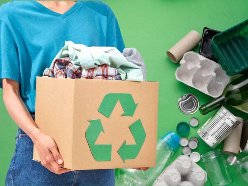 Osoba drži kartonsku kutiju sa reciklažnim znakom u kojoj se nalaze odeća i tekstil, okružena različitim materijalima za reciklažu poput plastike, stakla i metala, jasno izražena ekološka svest.