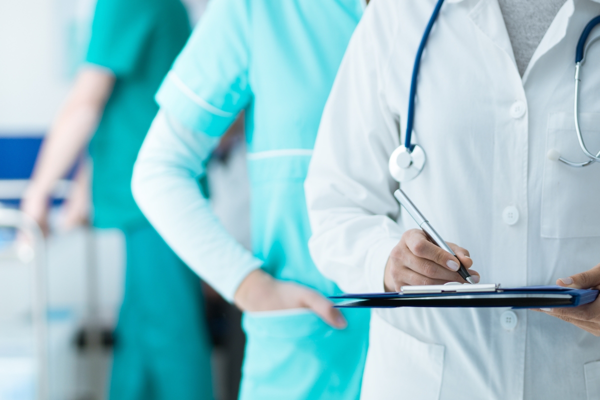 Medicinski radnik beleži informacije o pacijentu u bolničkom okruženju, dok zdravstveno osoblje stoji u pozadini.