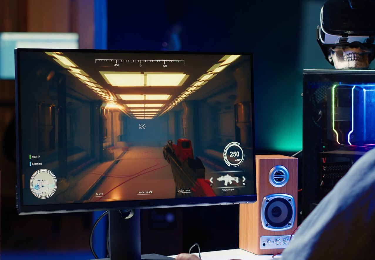 Gejming monitor sa prikazom pucačke video-igre, pored RGB računarskog kućišta i zvučnika na stolu