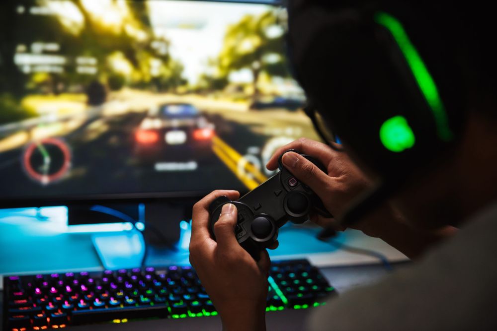 Gejmer sa slušalicama drži kontroler ispred monitora sa trkačkom igrom, dok se na stolu nalazi RGB tastatura